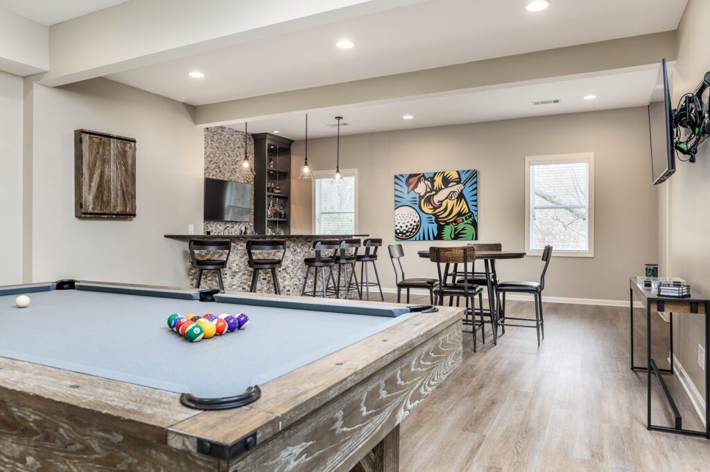 Basement game room featuring a pool table with colorful balls, modern bar area with seating, and vibrant wall art, showcasing a stylish and functional living space designed for entertainment and relaxation.