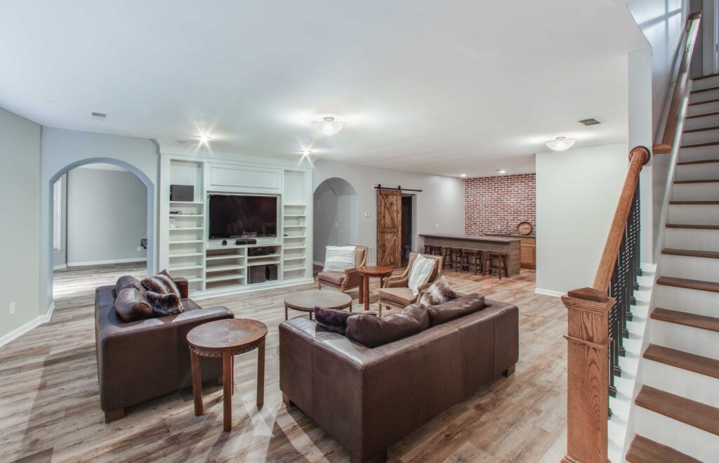 Modern basement living space with leather sofas, wooden coffee table, and open layout, featuring a media center and rustic brick accent wall, showcasing whole home remodeling design in Alpharetta, GA.
