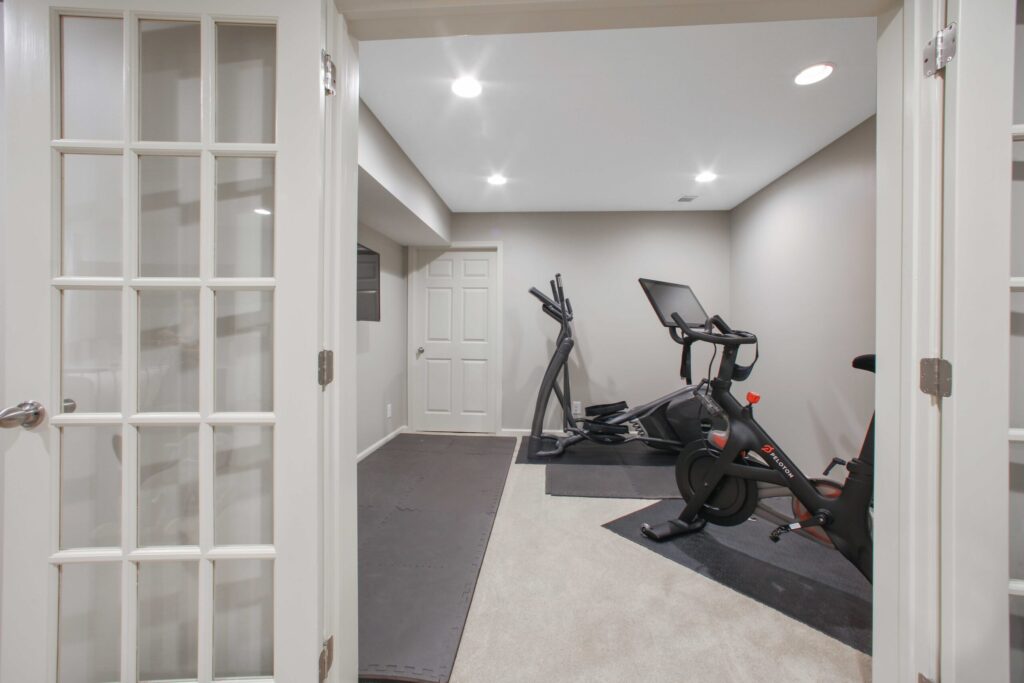 Basement home gym featuring exercise equipment, including an elliptical and stationary bike, with modern design elements and neutral color scheme, illustrating potential for functional home additions.