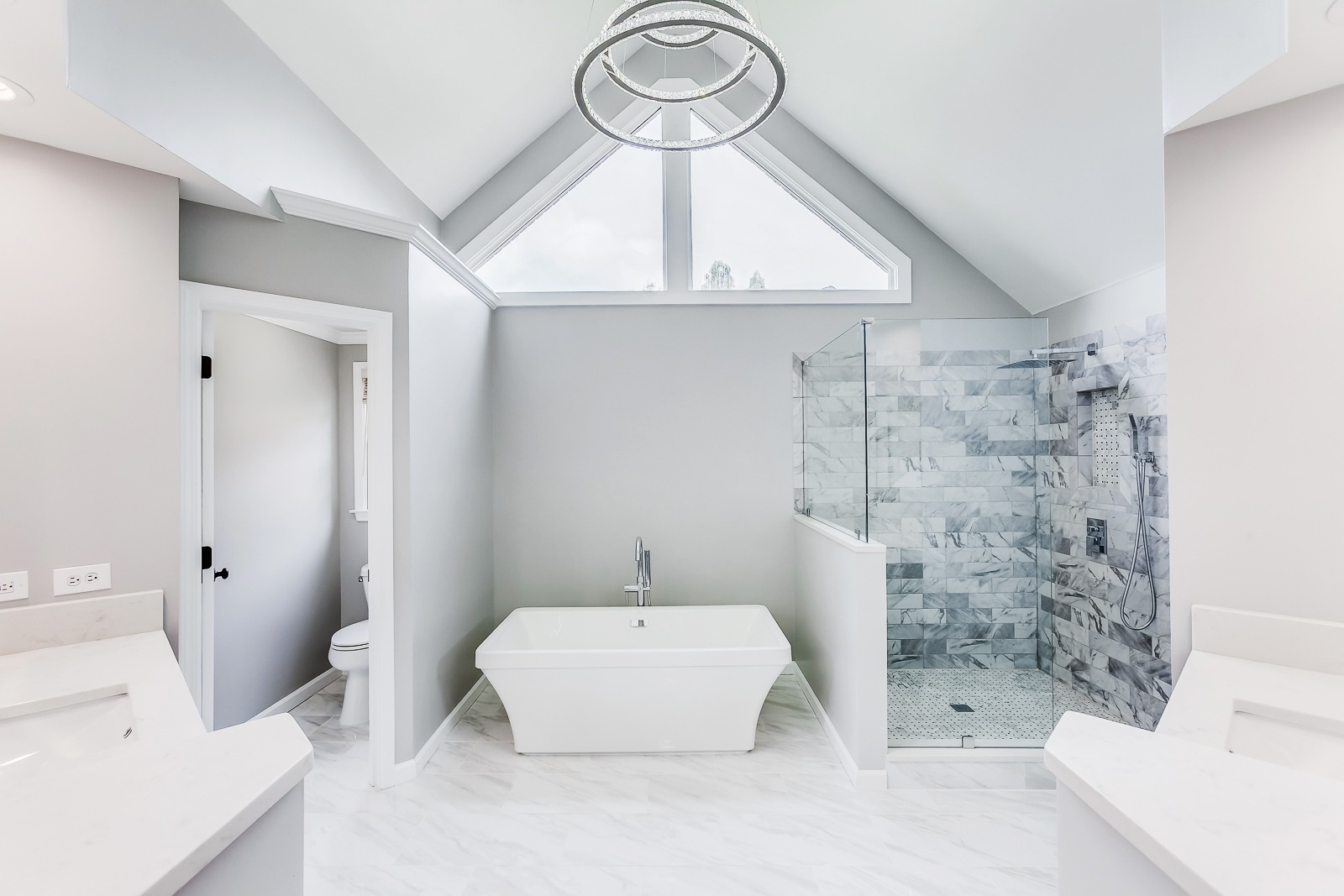 Modern bathroom featuring a freestanding white tub, frameless glass shower with marble tiles, and a high ceiling with a decorative chandelier, showcasing a clean and spacious design.