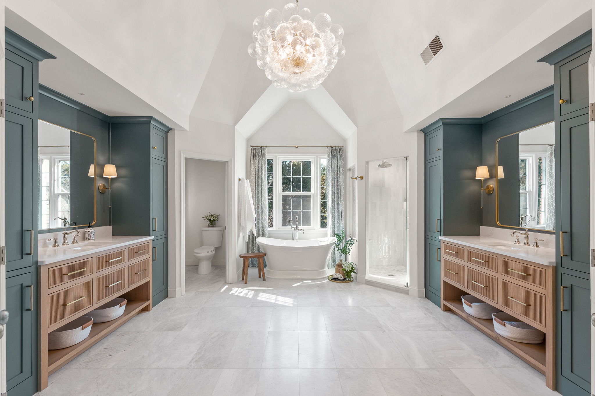 Luxurious bathroom renovation featuring dual vanities with wooden and blue cabinetry, a freestanding soaking tub, elegant chandelier, and glass-enclosed shower, showcasing HomeVantage's craftsmanship.
