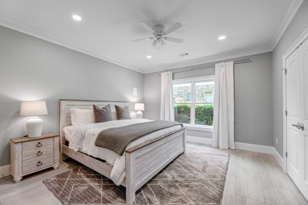 Modern bedroom interior featuring a light gray color scheme, wooden bed frame with decorative pillows, bedside table with lamp, and large window with white curtains, illustrating a comfortable living space ideal for home additions in Cumming, GA.