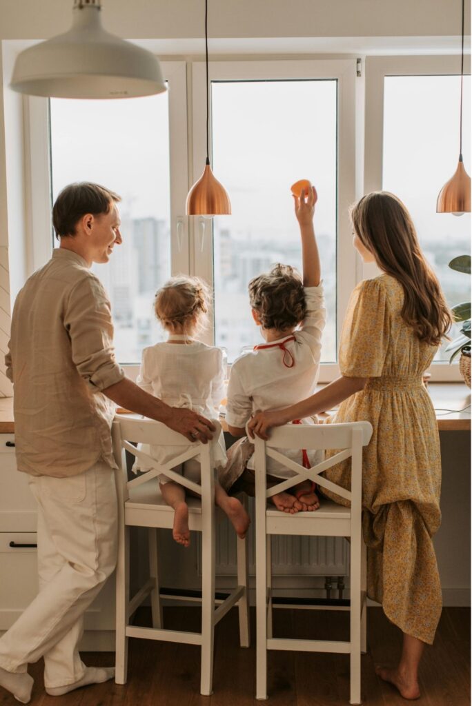 Family enjoying quality time in a modern kitchen, children seated on bar stools, one child raising an orange, emphasizing home life and family bonding.
