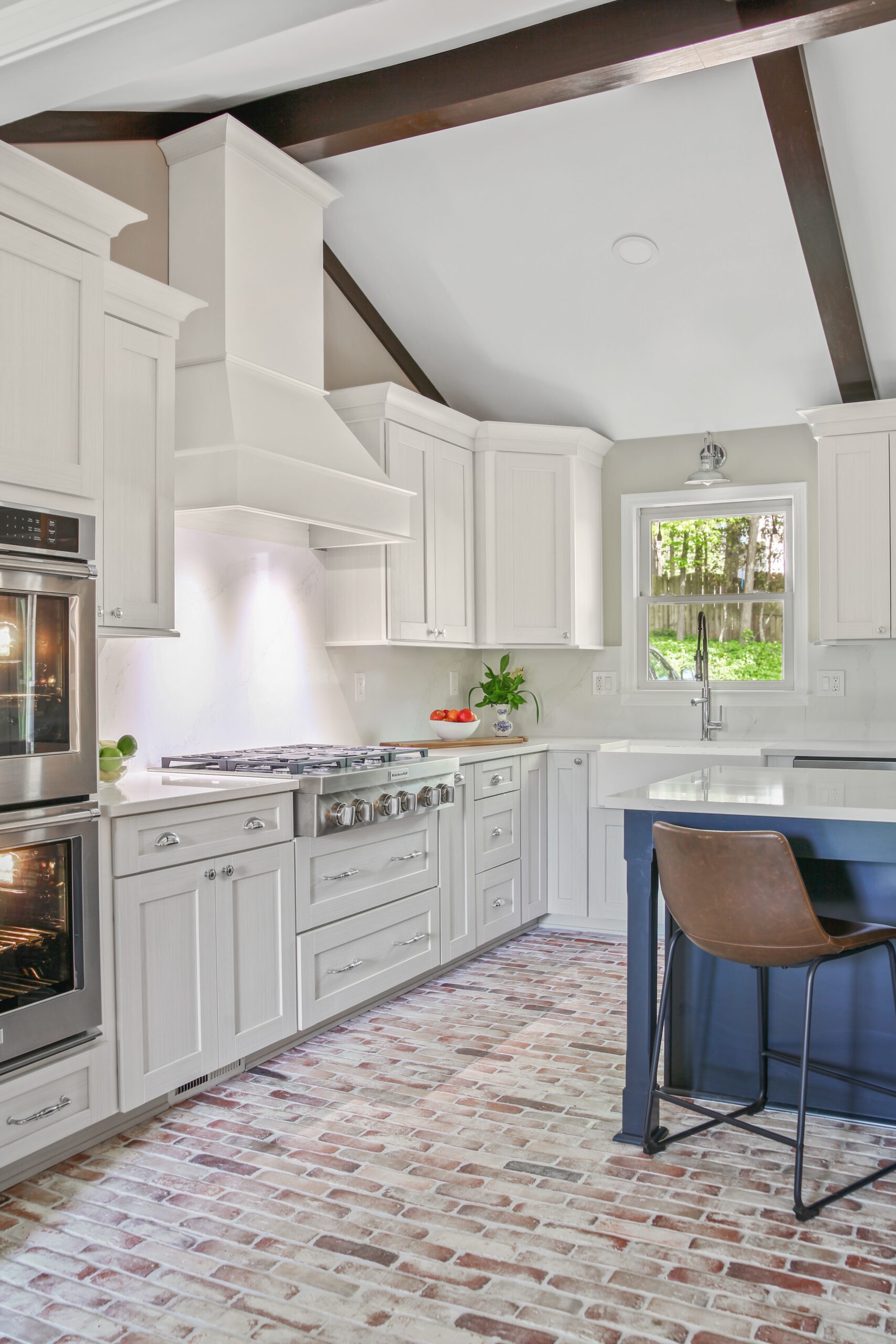 Modern kitchen design featuring white cabinetry, a stainless steel range, and a spacious island with a dark blue base, complemented by a brick-patterned floor and natural light from a nearby window.