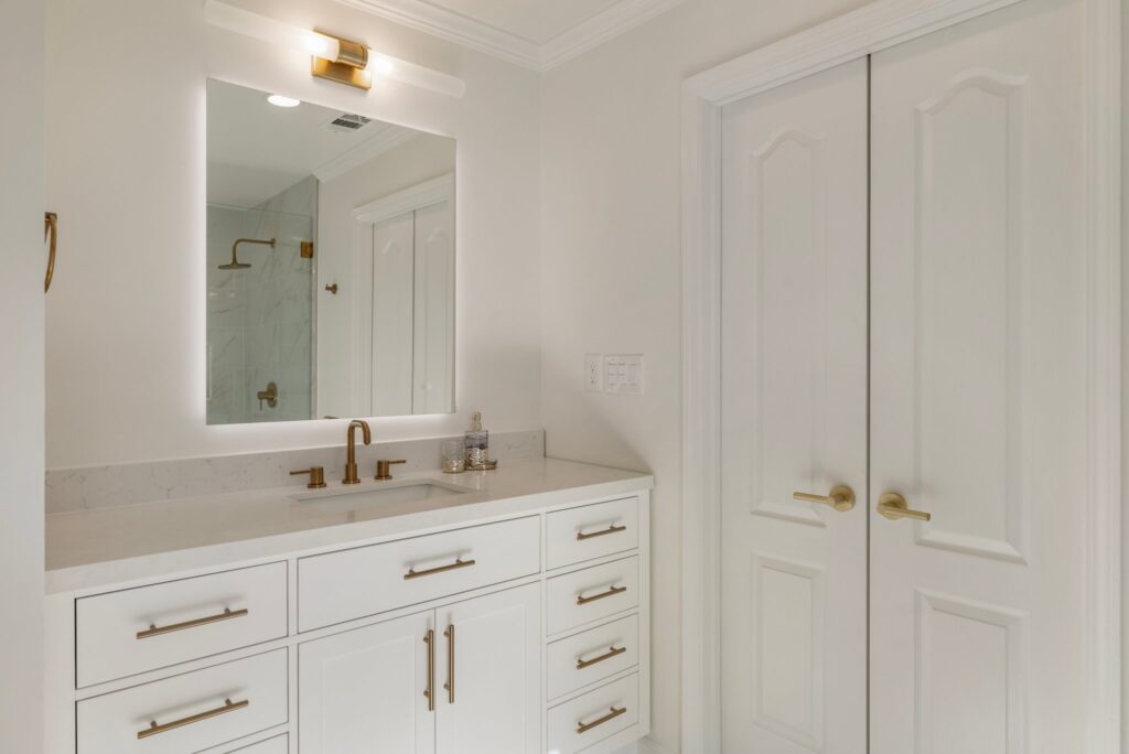 Modern bathroom with white cabinetry, gold fixtures, backlit mirror, and double doors in Park Brooke Trace remodeling project by HomeVantage.