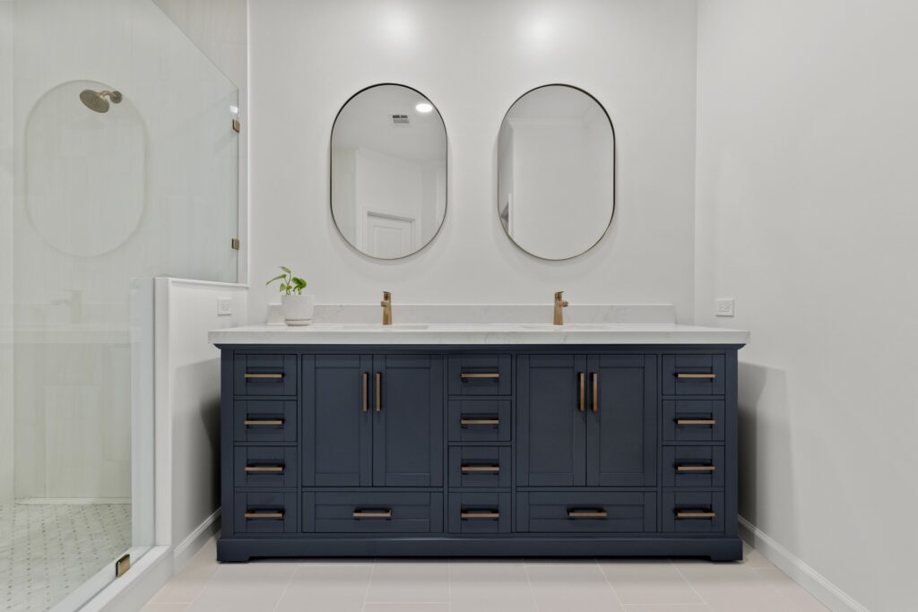 Modern bathroom renovation featuring a navy blue double vanity with gold hardware, two oval mirrors, and a glass shower enclosure, showcasing HomeVantage's design expertise in home improvement.