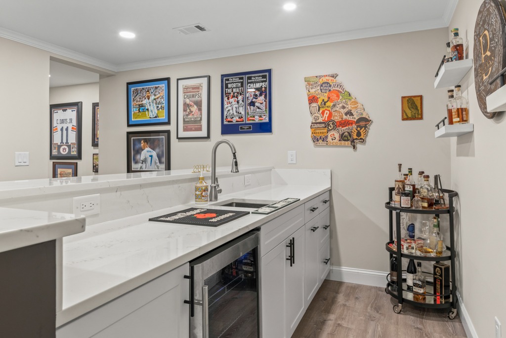 Modern basement bar area featuring white cabinetry, a sleek countertop, framed sports memorabilia, a Georgia state map art piece, and a cart displaying various liquor bottles, highlighting the Shepherds Pond remodeling project by HomeVantage in Cumming, GA.
