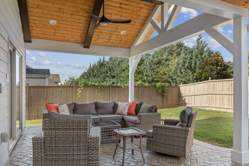 Outdoor living space featuring a covered patio with a sectional sofa, accent pillows, and a glass-top coffee table, surrounded by a fenced yard and greenery, showcasing HomeVantage's outdoor renovation expertise.