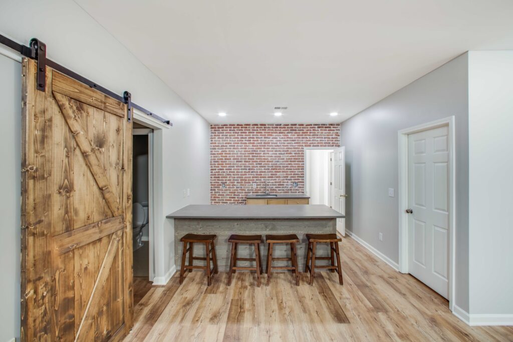 Basement remodeling project by HomeVantage featuring a modern kitchen with a concrete island, wooden bar stools, and a rustic sliding barn door against a brick accent wall in Roswell, GA.