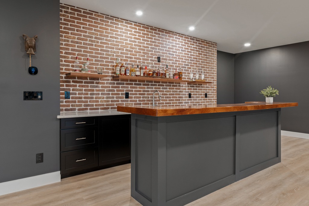 Modern basement bar area with exposed brick wall, wooden countertop, and stylish shelving displaying liquor bottles, showcasing the 'Wild Violet' remodeling project by HomeVantage in Cumming, GA.